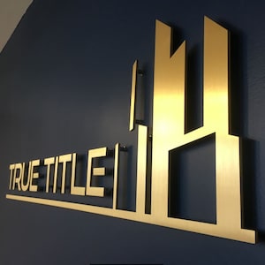 Pu&ograve; includere: Un cartello in metallo dorato con il testo "TRUE TITLE" e un design di edificio stilizzato.