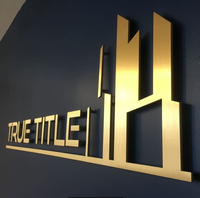 Pu&ograve; includere: Un cartello in metallo dorato con il testo "TRUE TITLE" e un design di edificio stilizzato.