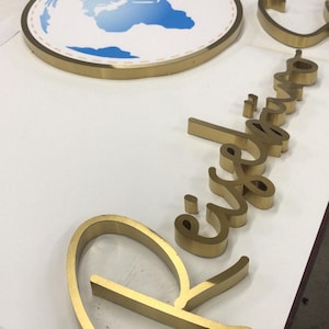 May include: A gold-toned metal sign featuring a circular emblem with a blue and white world map graphic and a small airplane icon. Beside it, gold-toned three-dimensional cursive lettering spells out the word Reiseb&uuml;ro in a script font.