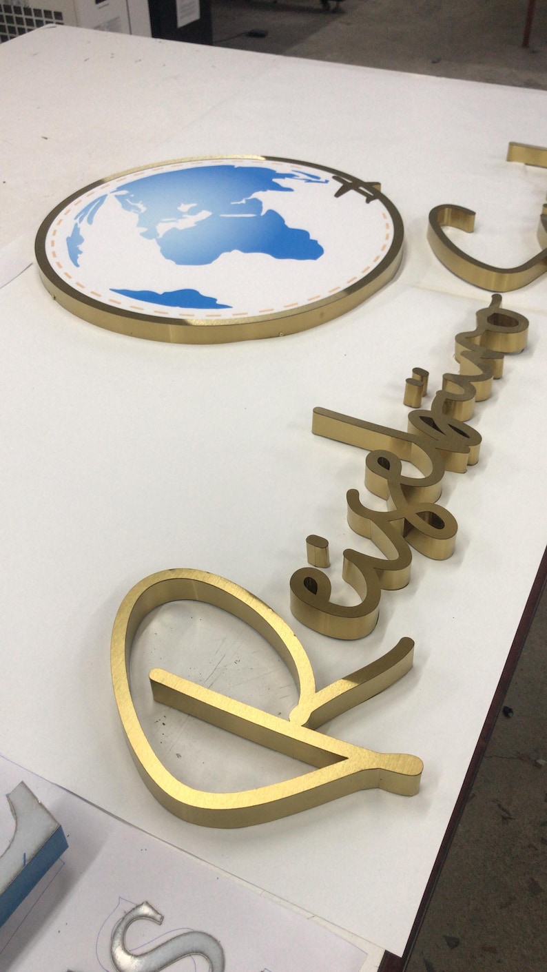 May include: A gold-toned metal sign featuring a circular emblem with a blue and white world map graphic and a small airplane icon. Beside it, gold-toned three-dimensional cursive lettering spells out the word Reiseb&uuml;ro in a script font.