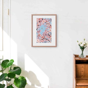 Op de afbeelding: Een ingelijste kunstdruk met een levendige illustratie van kersenbloesemtakken met roze bloemen tegen een lichtblauwe lucht. Het kunstwerk wordt gepresenteerd in een lichtbruine houten lijst.