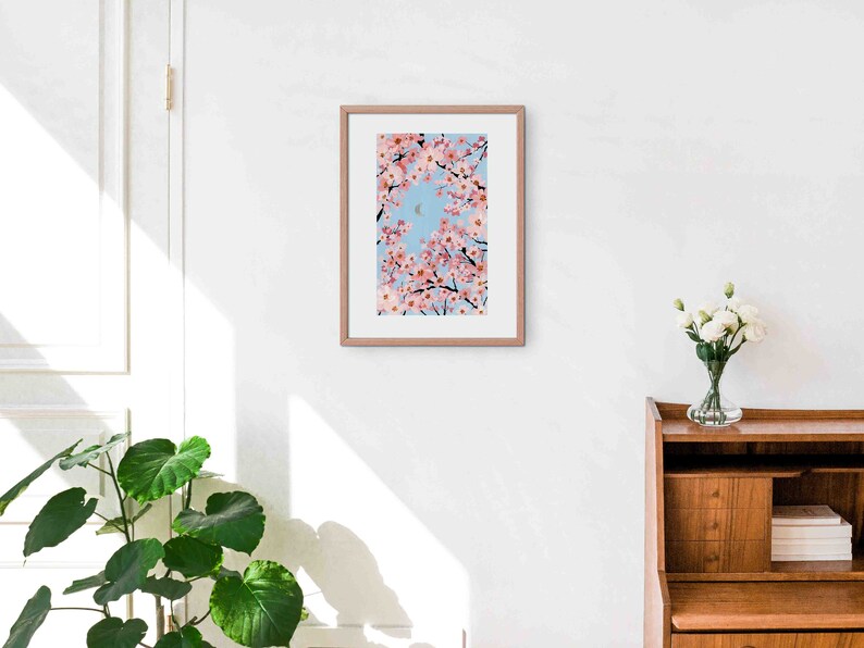 Op de afbeelding: Een ingelijste kunstdruk met een levendige illustratie van kersenbloesemtakken met roze bloemen tegen een lichtblauwe lucht. Het kunstwerk wordt gepresenteerd in een lichtbruine houten lijst.