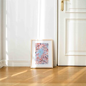 Op de afbeelding: Een ingelijste kunstdruk met roze kersenbloesems tegen een lichtblauwe lucht. Het kunstwerk is in een licht houten frame en staat op een houten vloer, leunend tegen een witte muur.