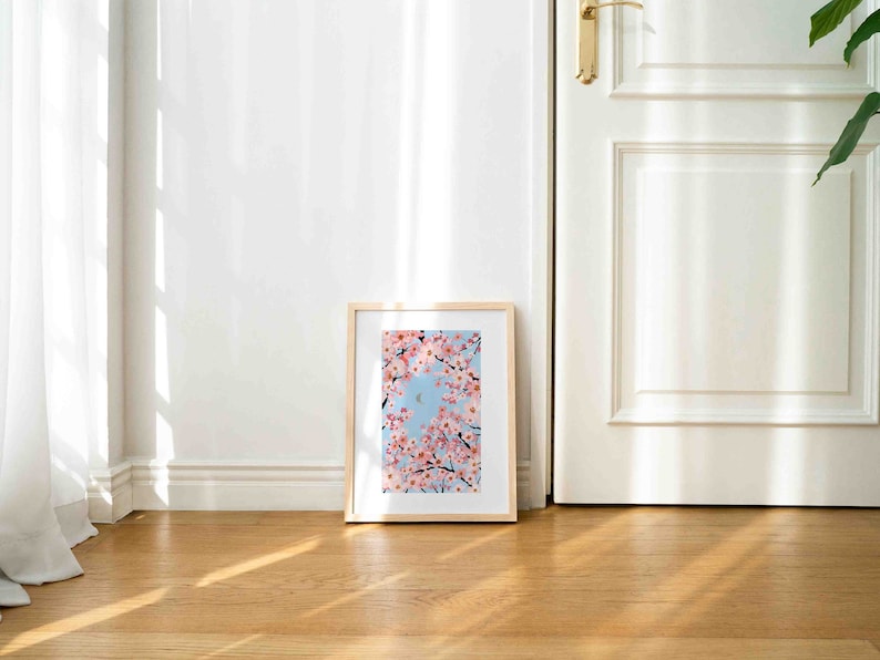 Op de afbeelding: Een ingelijste kunstdruk met roze kersenbloesems tegen een lichtblauwe lucht. Het kunstwerk is in een licht houten frame en staat op een houten vloer, leunend tegen een witte muur.