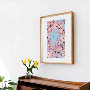 Op de afbeelding: Een ingelijste kunstdruk met een schilderij van roze kersenbloesems tegen een lichtblauwe lucht met een halve maan. Het kunstwerk zit in een houten lijst en hangt aan een witte muur boven een houten kast. Op de kast staat een vaas met gele tulpen.