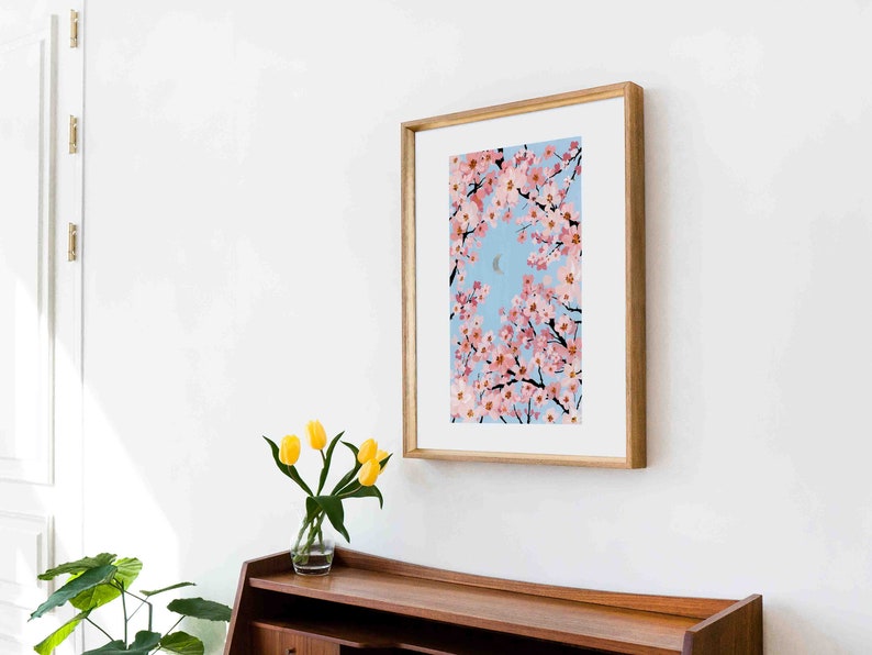 Op de afbeelding: Een ingelijste kunstdruk met een schilderij van roze kersenbloesems tegen een lichtblauwe lucht met een halve maan. Het kunstwerk zit in een houten lijst en hangt aan een witte muur boven een houten kast. Op de kast staat een vaas met gele tulpen.