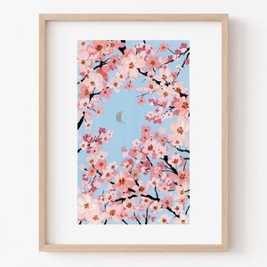 Op de afbeelding: Een ingelijste kunstdruk met een illustratie van kersenbloesemtakken met roze bloemen tegen een lichtblauwe lucht. Een zilveren halve maan is zichtbaar. Het kunstwerk is ingelijst in een licht houten frame.