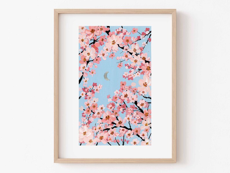 Op de afbeelding: Een ingelijste kunstdruk met een illustratie van kersenbloesemtakken met roze bloemen tegen een lichtblauwe lucht. Een zilveren halve maan is zichtbaar. Het kunstwerk is ingelijst in een licht houten frame.