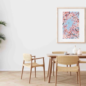 Op de afbeelding: Een ingelijste kunstdruk toont kersenbloesemtakken tegen een lichtblauwe lucht met een halve maan. De print zit in een licht houten frame. De sc&egrave;ne speelt zich af in een eetkamer met een houten tafel en gevlochten stoelen.