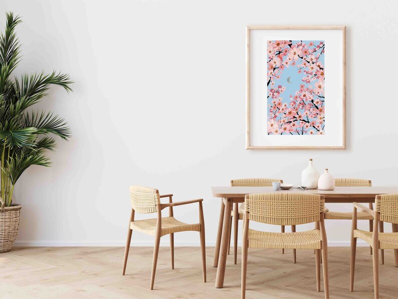 Op de afbeelding: Een ingelijste kunstdruk toont kersenbloesemtakken tegen een lichtblauwe lucht met een halve maan. De print zit in een licht houten frame. De sc&egrave;ne speelt zich af in een eetkamer met een houten tafel en gevlochten stoelen.
