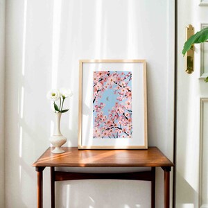 Op de afbeelding: Een ingelijste kunstdruk met kersenbloesemtakken tegen een lichtblauwe lucht, met een halve maan. Het kunstwerk wordt tentoongesteld op een houten tafel naast een witte vaas met witte bloemen.