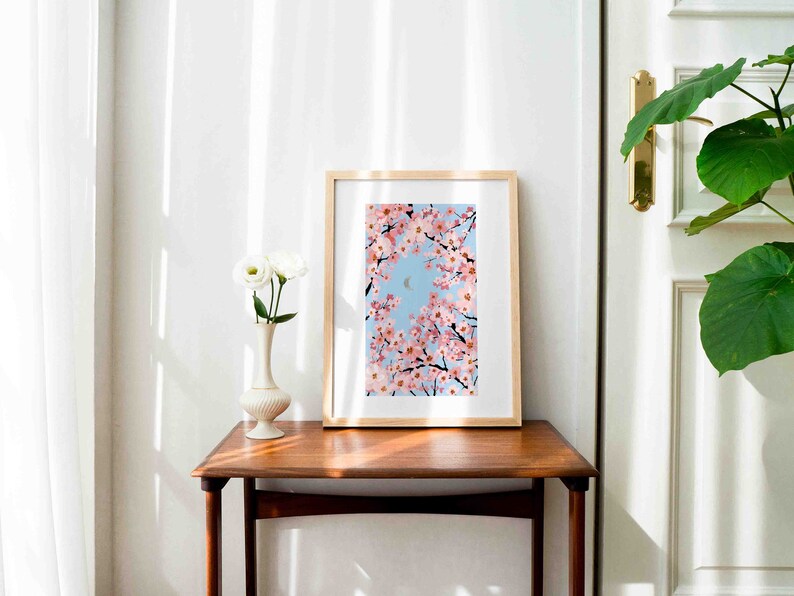 Op de afbeelding: Een ingelijste kunstdruk met kersenbloesemtakken tegen een lichtblauwe lucht, met een halve maan. Het kunstwerk wordt tentoongesteld op een houten tafel naast een witte vaas met witte bloemen.