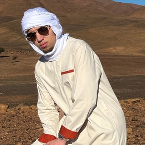 Puede incluir: Una persona con una túnica color crema con ribetes naranjas y un turbante blanco. La persona también lleva gafas de sol. El fondo presenta un paisaje montañoso con un cielo azul claro.