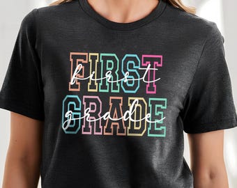 May include: A charcoal grey short-sleeve t-shirt featuring the text "FIRST GRADE" in a blocky, multi-coloured varsity font. Overlaid on top of the block letters is the word "first grade" written in a white, flowing cursive script font.