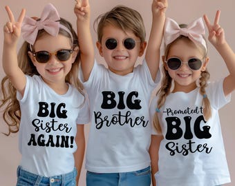 May include: Three white t-shirts with black text. The left shirt reads BIG SISTER AGAIN!, the center shirt reads BIG Brother, and the right shirt reads Promoted to BIG Sister. The shirts are worn with blue denim jeans and round sunglasses.