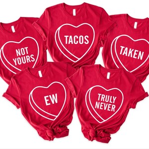 May include: Red t-shirts with white heart-shaped graphics. Each heart contains a different phrase: "NOT YOURS", "TACOS", "TAKEN", "EW", and "TRULY NEVER". The shirts are casual wear.