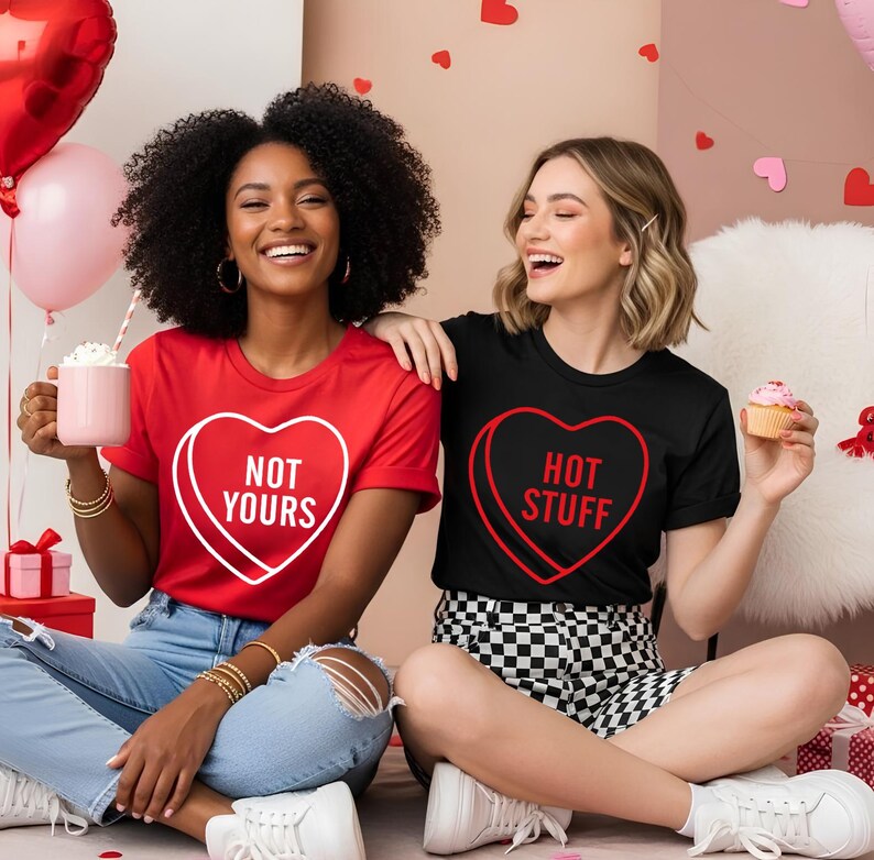May include: Two women wearing matching Valentine's Day t-shirts. One shirt is red with a white heart that says "NOT YOURS". The other shirt is black with a red heart that says "HOT STUFF". Both women are smiling.