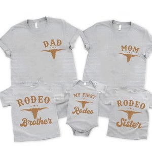 May include: A collection of five light gray heather t-shirts and one baby bodysuit featuring brown screen-printed graphics. The shirts display the text DAD, MOM, RODEO BROTHER, and RODEO SISTER, while the bodysuit reads MY FIRST RODEO.