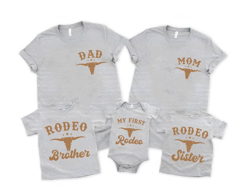 May include: A collection of five light gray heather t-shirts and one baby bodysuit featuring brown screen-printed graphics. The shirts display the text DAD, MOM, RODEO BROTHER, and RODEO SISTER, while the bodysuit reads MY FIRST RODEO.
