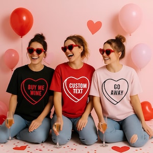 May include: Three people wearing heart-shaped sunglasses and t-shirts with candy heart designs. The shirts are black, red, and pink, with phrases like "BUY ME WINE," "CUSTOM TEXT," and "GO AWAY." Balloons and heart decorations complete the Valentine's Day theme.