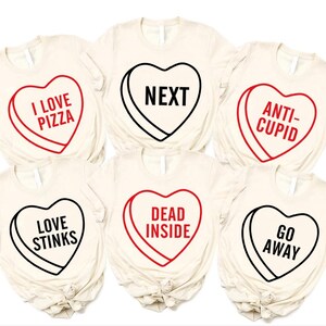 May include: Six cream-colored t-shirts with heart-shaped candy graphics. Each heart has a different phrase: "I LOVE PIZZA," "NEXT," "ANTI-CUPID," "LOVE STINKS," "DEAD INSIDE," and "GO AWAY." The hearts are outlined in black or red.