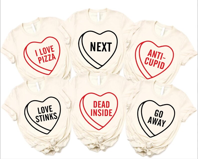 May include: Six cream-colored t-shirts with heart-shaped candy graphics. Each heart has a different phrase: "I LOVE PIZZA," "NEXT," "ANTI-CUPID," "LOVE STINKS," "DEAD INSIDE," and "GO AWAY." The hearts are outlined in black or red.