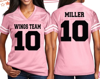 Puede incluir: Camiseta de fútbol rosa con rayas blancas en las mangas. La parte delantera de la camiseta tiene el texto "WINGS TEAM" y el número "10" en negro. La parte trasera de la camiseta tiene el nombre "MILLER" y el número "10" en negro.