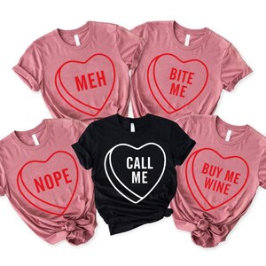 May include: Five t-shirts with heart-shaped candy designs. Four are pink with red text: "MEH", "BITE ME", "NOPE", and "BUY ME WINE". The fifth is black with white text: "CALL ME".