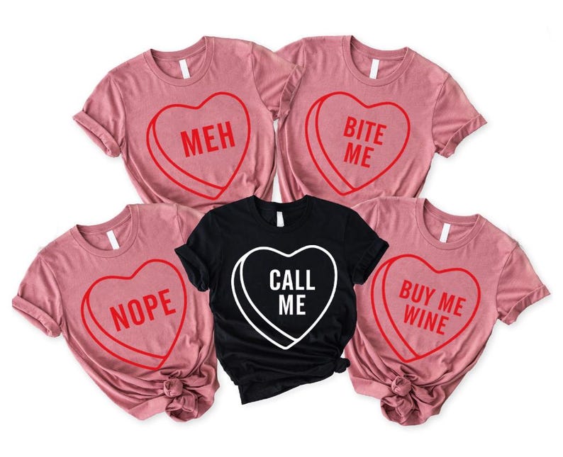 May include: Five t-shirts with heart-shaped candy designs. Four are pink with red text: "MEH", "BITE ME", "NOPE", and "BUY ME WINE". The fifth is black with white text: "CALL ME".