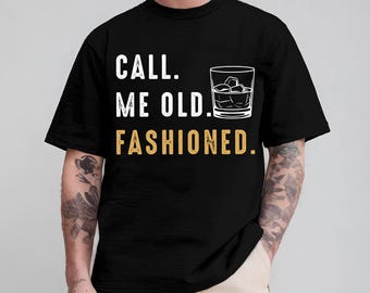 May include: A black short-sleeve t-shirt featuring the white text "CALL. ME OLD." above a graphic of a glass filled with ice cubes. Below the glass, the word "FASHIONED." is printed in a tan color. The design has a distressed texture.