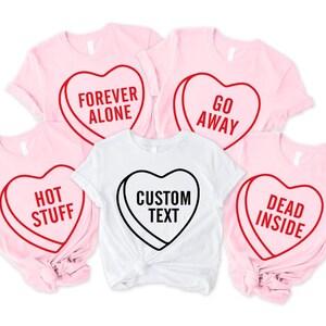 May include: A collection of pink and white t-shirts with heart-shaped designs. The pink shirts have red outlines with phrases like "Forever Alone," "Go Away," "Hot Stuff," and "Dead Inside." The white shirt features "Custom Text."