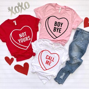 May include: Three t-shirts with heart-shaped candy graphics and text. A red shirt says "NOT YOURS," a pink shirt says "BOY BYE," and a white shirt says "CALL ME." A pair of blue jeans and decorative hearts are also in the image.