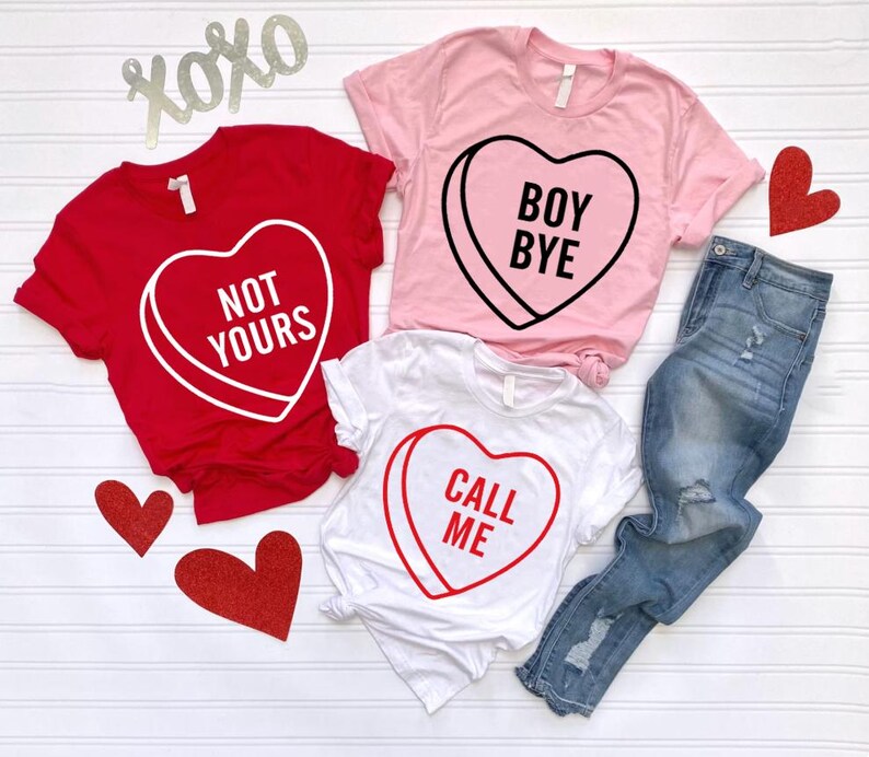 May include: Three t-shirts with heart-shaped candy graphics and text. A red shirt says "NOT YOURS," a pink shirt says "BOY BYE," and a white shirt says "CALL ME." A pair of blue jeans and decorative hearts are also in the image.