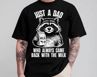 May include: A black short-sleeved t-shirt featuring a white graphic of a raccoon holding a milk carton. The text above reads JUST A DAD and the text below reads WHO ALWAYS CAME BACK WITH THE MILK. The design is centered on the chest of the garment.
