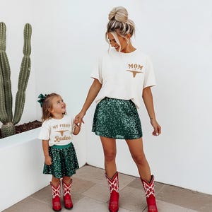 May include: Two off-white t-shirts with brown graphics. The adult shirt features the text MOM with a longhorn skull. The child shirt features the text MY FIRST Rodeo with a longhorn skull. Both are paired with dark green sequin skirts and red boots.