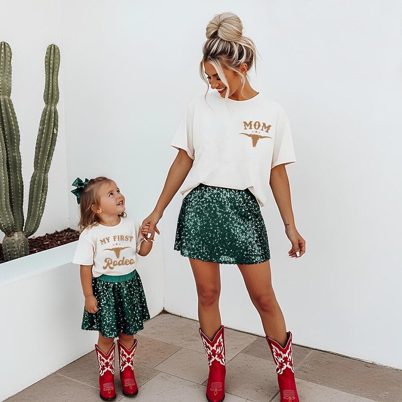 May include: Two off-white t-shirts with brown graphics. The adult shirt features the text MOM with a longhorn skull. The child shirt features the text MY FIRST Rodeo with a longhorn skull. Both are paired with dark green sequin skirts and red boots.