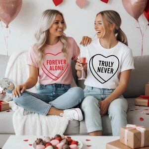 May include: Two women wearing t-shirts with candy heart designs. One shirt is pink with "FOREVER ALONE" in red, the other is white with "TRULY NEVER" in black. Both are seated on a couch, with balloons and gifts in the background.