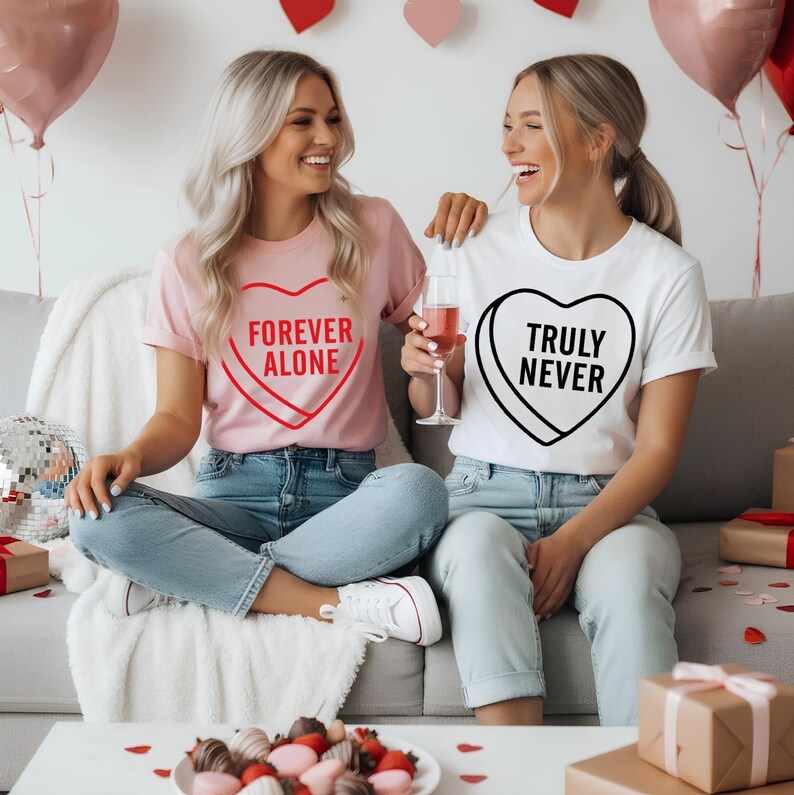 May include: Two women wearing t-shirts with candy heart designs. One shirt is pink with "FOREVER ALONE" in red, the other is white with "TRULY NEVER" in black. Both are seated on a couch, with balloons and gifts in the background.