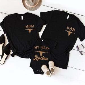 May include: Two black short-sleeved t-shirts and one black infant bodysuit. The t-shirts feature a tan graphic of a bull skull and the text MOM or DAD. The bodysuit features a tan bull skull graphic and the text MY FIRST RODEO.