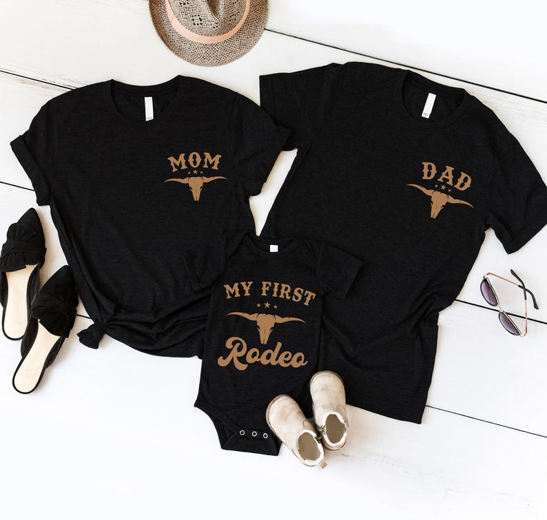 May include: Two black short-sleeved t-shirts and one black infant bodysuit. The t-shirts feature a tan graphic of a bull skull and the text MOM or DAD. The bodysuit features a tan bull skull graphic and the text MY FIRST RODEO.