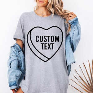May include: A gray t-shirt featuring a black heart-shaped candy design with the words "CUSTOM TEXT" in bold, black letters. The person is wearing a light blue denim jacket over the t-shirt.
