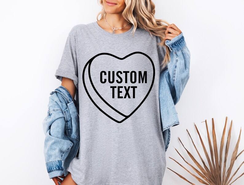 May include: A gray t-shirt featuring a black heart-shaped candy design with the words "CUSTOM TEXT" in bold, black letters. The person is wearing a light blue denim jacket over the t-shirt.
