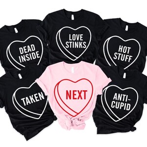 May include: A collection of black and pink t-shirts with heart-shaped candy designs. Each shirt features a different phrase in white or red, including "DEAD INSIDE," "LOVE STINKS," "HOT STUFF," "TAKEN," "NEXT," and "ANTI-CUPID."