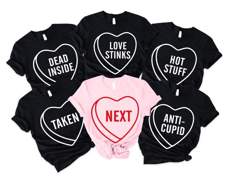May include: A collection of black and pink t-shirts with heart-shaped candy designs. Each shirt features a different phrase in white or red, including "DEAD INSIDE," "LOVE STINKS," "HOT STUFF," "TAKEN," "NEXT," and "ANTI-CUPID."