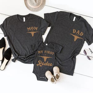 May include: Two dark heather gray short-sleeve t-shirts and one matching infant bodysuit. The shirts feature the text MOM or DAD above a tan bull skull graphic. The bodysuit displays the text MY FIRST RODEO above the same bull skull graphic.