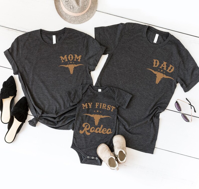 May include: Two dark heather gray short-sleeve t-shirts and one matching infant bodysuit. The shirts feature the text MOM or DAD above a tan bull skull graphic. The bodysuit displays the text MY FIRST RODEO above the same bull skull graphic.