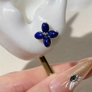 Peut inclure: Une vue rapprochée d'une boucle d'oreille en forme de fleur bleue à cinq pétales. La boucle d'oreille est en argent et a une petite pierre transparente au centre de chaque pétale.