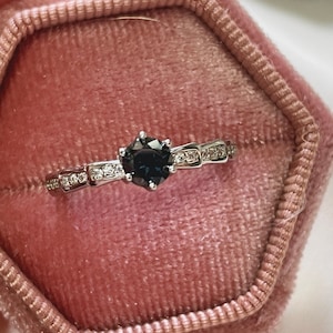 Puede incluir: Un anillo de compromiso de diamante negro con una banda de plata y pequeños diamantes engastados en la banda. El anillo está en una caja de anillo de terciopelo rosa.