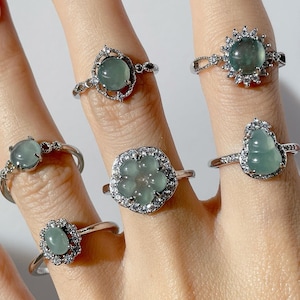 Peut inclure: Cinq bagues en argent avec des pierres de jade vert serties dans différents styles. Les bagues sont portées sur une main avec un vernis à ongles blanc.