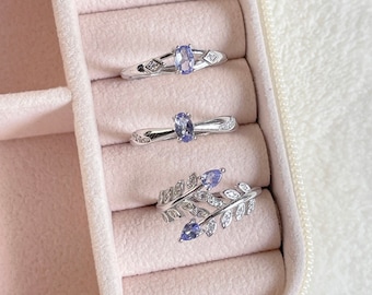 May include: Three silver rings with blue gemstones and small clear stones. The rings are displayed in a pink velvet ring box.